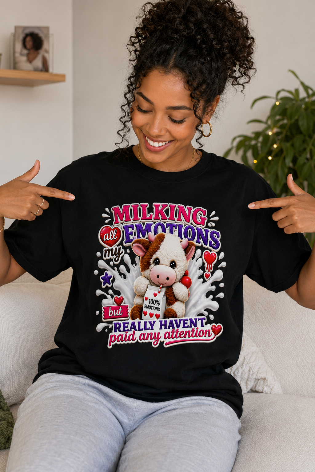 Woman smiling and pointing to black graphic t-shirt with cute cow design that reads "Milking My Emotions But Really Haven't Paid Any Attention"